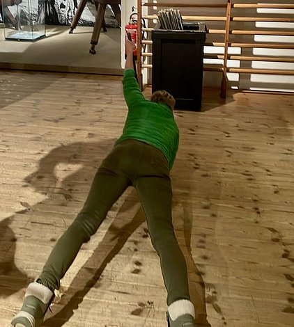 Eine Person liegt am Boden und streckt sich mit einer Hand nach einem Objekt. Sie trägt ein grünes Oberteil und graue Hose. Der Boden ist aus Holz. Im Hintergrund sind schemenhaft einige Möbel und andere Objekte zu erkennen.