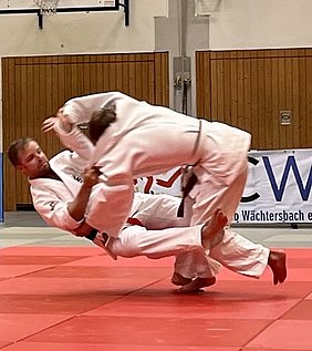 Zwei Judokas in weißen Anzügen führen eine Technik im Wettkampf aus. Einer der Kämpfer hebt den anderen an, während dieser sich in der Luft befindet. Der Boden ist mit einer roten Judomatte bedeckt.