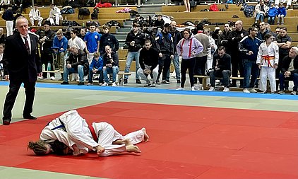 Ein Judo-Wettkampf auf der Matte, zwei Kämpfer in weißen Judogis. Einer der Kämpfer, mit rotem Gürtel, liegt auf dem Rücken, während der andere ihn hält. Im Hintergrund sind Zuschauer auf Bänken zu sehen, die das Geschehen aufmerksam verfolgen. Ein Schiedsrichter steht am Rand der Matte.