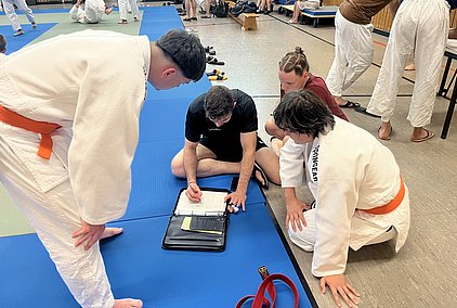 Vier Judokas in Trainingsanzügen stehen oder sitzen um einen Tisch, während einer von ihnen in einem Notizbuch schreibt. Die Athleten scheinen sich über das Training oder eine Strategie auszutauschen. Der Boden ist mit blauen und grünen Matten bedeckt, die Umgebung wirkt sportlich und aktiv.
