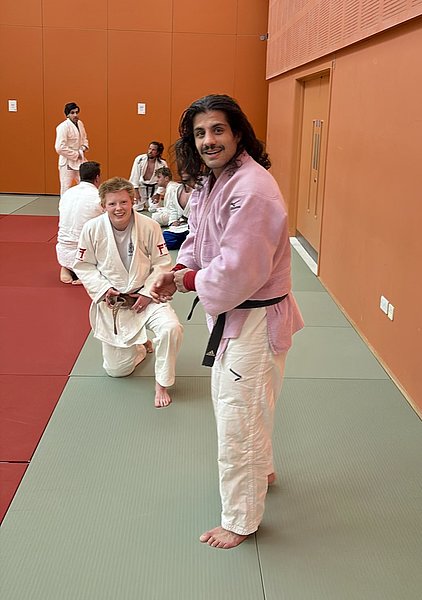 Auf der Matte sind mehrere Judoka in weißen und einem pinken Gi. Ein Mann in pinkem Gi steht lachend und interagiert mit einem anderen, der im Knien sitzt und einen braunen Gürtel trägt. Der Hintergrund ist orange, und die Matte ist in grünen und roten Bereichen unterteilt.