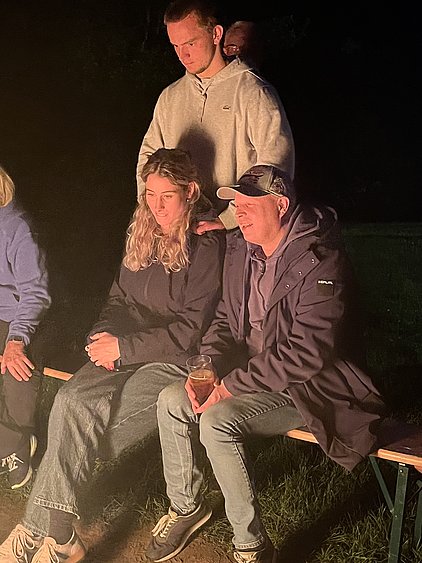 Eine Gruppe von Menschen sitzt um ein Lagerfeuer. Ein Mann steht hinter einer Frau mit langen, lockigen Haaren, die neben einem weiteren Mann sitzt, der ein Getränk in der Hand hält. Die Szene ist in der Dunkelheit beleuchtet, was eine gemütliche Stimmung vermittelt.