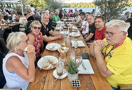 Eine Gruppe von acht Personen sitzt an einem Holztisch draußen, mit leeren Tellern und Getränken vor sich. Sie lächeln und scheinen eine angenehme Zeit zusammen zu verbringen. Im Hintergrund ist ein schöner Blick auf Wasser und Bäume zu sehen.