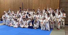 Eine Gruppe von Kindern in Judoanzügen posiert auf einer blauen Matte. Sie halten Urkunden in den Händen und tragen Medaillen. Die Kinder zeigen stolz ihre erhobenen Fäuste, was auf Freude und Erfolge bei einem Wettkampf hinweist. Der Hintergrund ist eine Holzwand in einer Turnhalle.