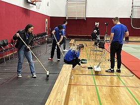 Eine Gruppe von fünf Personen in blauen T-Shirts arbeitet gemeinsam in einer Sporthalle. Sie reinigen den Boden mit Besen und Mop, während einige auf einer Sitzbank warten. Im Hintergrund sind Sportmatten sichtbar, und ein Kamerateam filmt die Aktivitäten.