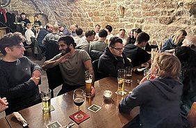 In einem gemütlichen Restaurant sitzen mehrere Gruppen an Tischen. Zwei Männer auf der linken Seite unterhalten sich, während andere Gäste Getränke vor sich haben. Die Wände sind aus Stein, und die Atmosphäre wirkt gesellig und lebhaft.