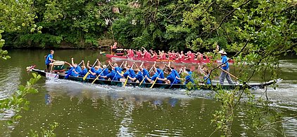 Zwei Drachenboote fahren auf einem ruhigen Gewässer. Die Mannschaft im vorderen Boot trägt blaue Shirts und rudert synchron, während ein Trommler die Rhythmen schlägt. Im hinteren Boot sind Teilnehmer in roten Shirts zu sehen, die ebenfalls rudern. Die Umgebung ist von Bäumen umgeben.