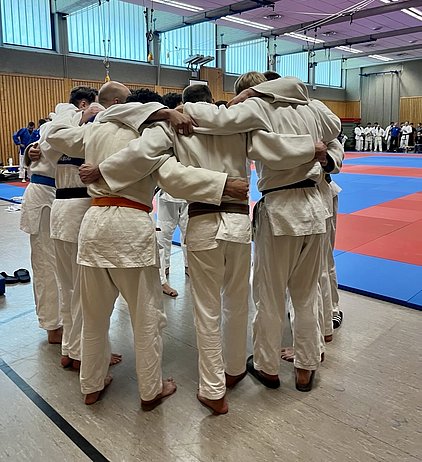 Eine Gruppe von Judokas steht eng umschlungen in einem Trainingsraum. Sie tragen traditionelle Judo-Kleidung und scheinen sich auf einen Wettkampf oder eine Übung vorzubereiten. Der Boden ist mit blauen und roten Matten ausgelegt, die Atmosphäre wirkt teamorientiert und fokussiert.