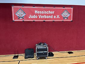 Ein rotes Banner mit der Aufschrift „Hessischer Judo Verband e.V.“ hängt an einer roten Wand. Darunter steht ein Mischpult mit verschiedenen angeschlossenen Geräten. Im Vordergrund sind einige Technikgegenstände sichtbar, die zur Beschallung verwendet werden können.