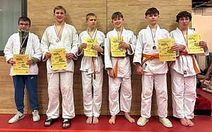 Sechs junge Judoka stehen in einem Reihen neben einander, tragen weiße Judogis mit orangefarbenen Gürteln und Medaillen um den Hals. Sie halten Urkunden in den Händen, die sie für ihre Leistungen bei einem Wettkampf erhalten haben. Der Hintergrund ist eine Holzwand in einem Sportraum.