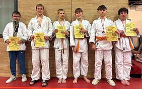 Sechs junge Judoka stehen in einem Reihen neben einander, tragen weiße Judogis mit orangefarbenen Gürteln und Medaillen um den Hals. Sie halten Urkunden in den Händen, die sie für ihre Leistungen bei einem Wettkampf erhalten haben. Der Hintergrund ist eine Holzwand in einem Sportraum.