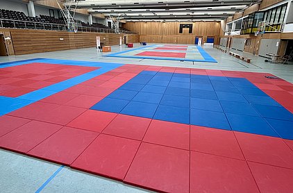 Die Aufnahme zeigt eine Sporthalle mit Matten in Rot und Blau. Die Matten sind in rechteckigen Bereichen angeordnet, die für eine Sportveranstaltung oder das Training vorbereitet sind. Im Hintergrund sind einige Sitzplätze und ein Basketballkorb sichtbar.