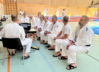 Eine Gruppe von Männern in Judoanzügen sitzt auf einer Bank und sieht in Richtung eines Laptops. Der Laptop steht auf einem Tisch. Im Hintergrund sind weitere Judoka zu sehen, die sich auf Matten vorbereiten. Die Sporthalle ist hell und hat Holzwände.