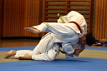 Zwei Judoka in traditionellen weißen Gi-Kleidung kämpfen auf einer Matte. Einer führt einen Wurf aus, während der andere sich verteidigt. Die Matte hat verschiedene Farben, und die Umgebung zeigt Holzwände, die auf einen Übungsraum hinweisen.