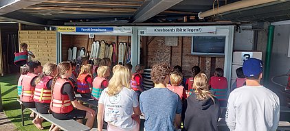Eine Gruppe von Kindern und Jugendlichen in Schwimmwesten sitzt auf Bänken und schaut auf einen Bildschirm, der eine Wasseraktivität zeigt. Im Hintergrund sind verschiedene Wassersportgeräte aufbewahrt. Die Szene findet in einem geschützten Bereich statt.
