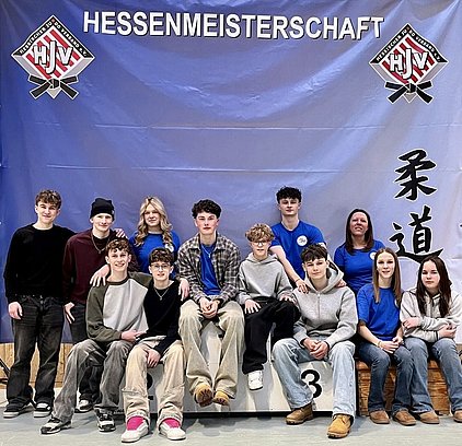 Eine Gruppe von zwölf Jugendlichen und Erwachsenen sitzt auf einer Bühne vor einem Banner mit der Aufschrift „Hessenmeisterschaft“. Sie tragen überwiegend blaue T-Shirts und lächeln in die Kamera. Im Hintergrund sind Kalligrafiezeichen sichtbar.