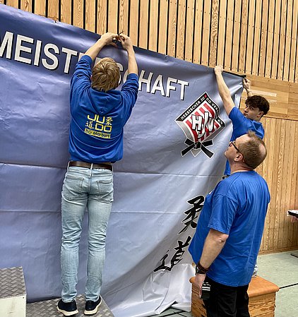 Drei Personen in blauen T-Shirts hängen ein großes Banner mit dem Wort "MEISTERSCHAFT" und einem Judo-Logo auf. Sie sind in einer Sporthalle und scheinen mit der Anbringung des Banners beschäftigt zu sein.
