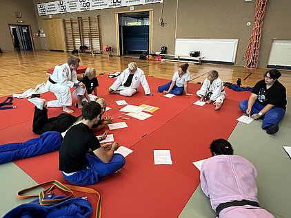 Eine Gruppe von Personen sitzt und liegt auf einer roten Mattenfläche in einem Sportraum. Einige tragen Judokleidung. Sie arbeiten an Aufgaben, schreiben Notizen und tauschen sich aus. Der Fokus liegt auf dem gemeinsamen Lernen und der Interaktion.