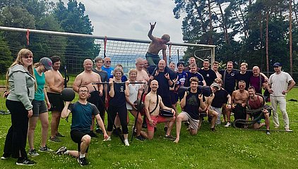 Eine Gruppe von etwa 30 Personen steht auf einer Wiese, viele sind oberkörperfrei und halten Fitnessgeräte wie Gewichte und Seile. Sie zeigen Freude und posieren vor einem Fußballtor, umgeben von Bäumen und einer bewölkten Himmel.