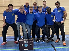 Eine Gruppe von acht Personen in blauer Judo-Bekleidung posiert lächelnd für ein Gruppenfoto. Sie stehen vor einer Holzwand in einer Turnhalle. Im Vordergrund ist ein Judo-Koffer mit einem Aufdruck sichtbar. Die Atmosphäre wirkt freundlich und sportlich.