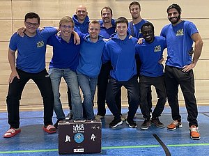 Eine Gruppe von acht Personen in blauer Judo-Bekleidung posiert lächelnd für ein Gruppenfoto. Sie stehen vor einer Holzwand in einer Turnhalle. Im Vordergrund ist ein Judo-Koffer mit einem Aufdruck sichtbar. Die Atmosphäre wirkt freundlich und sportlich.