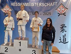 Eine Gruppe junger Judoka steht auf einem Treppchen bei der Hessenmeisterschaft. Die ersten drei Platzierungen sind sichtbar, die Sportlerinnen tragen Medaillen und halten Urkunden. Im Vordergrund stehen zwei Erwachsene und im Hintergrund ein Banner mit dem Schriftzug "Hessenmeisterschaft".