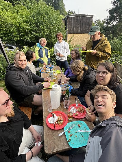 Eine Gruppe von Menschen sitzt an einem Holztisch im Freien und genießt ein Essen. Auf den Tellern sind Reste von verschiedenen Gerichten zu sehen. Einige Personen lachen und unterhalten sich, während andere mit Essen beschäftigt sind. Im Hintergrund sieht man viel grünes Laub.