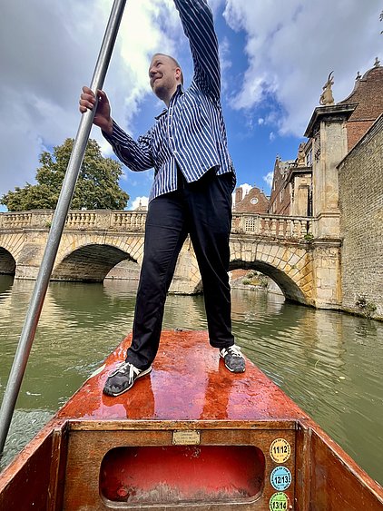 Ein Mann steht auf einem roten Boot und lenkt mit einem langen Stab. Im Hintergrund ist eine Steinbrücke und Bäume zu sehen, der Himmel ist teilweise bewölkt. Der Mann trägt ein gestreiftes Hemd und schwarze Hosen.