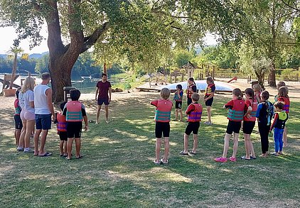 Eine Gruppe von Kindern in Schwimmwesten steht auf einer Wiese und hört einem Trainer zu, der vor ihnen steht. Im Hintergrund ist ein Gewässer zu sehen, umgeben von Bäumen und Freizeitmöglichkeiten.