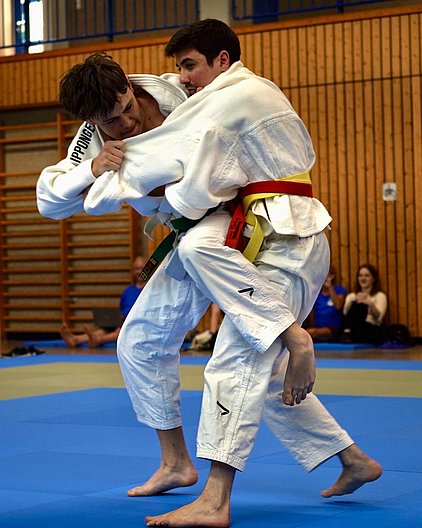 Zwei Judokas im Wettkampf auf einer blauen Matte. Der Judo-ka mit der grünen und gelben Schärpe versucht, seinen Gegner zu überwältigen. Die Umgebung zeigt eine Sporthalle mit Zuschauern im Hintergrund.