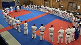 Eine Gruppe von Judoka steht in einem Kreis auf einer Kampfsportmatte. Die Teilnehmer tragen weiße und schwarze Judogis und applaudieren. In der Mitte sind einige Trainer, die sich mit den Schülern austauschen. Der Raum hat eine sportliche Atmosphäre.