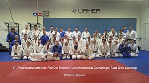 Eine Gruppe von etwa 50 Personen in Judo-Anzügen steht auf einer Judomatte, lächelnd und in verschiedenen Posen. Die Szene findet bei einem Freundschaftstreffen der Judovereine FinnDai Helsinki, Universität Cambridge und Blau-Gelb Marburg in Helsinki 2024 statt.