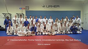 Eine Gruppe von etwa 50 Personen in Judo-Anzügen steht auf einer Judomatte, lächelnd und in verschiedenen Posen. Die Szene findet bei einem Freundschaftstreffen der Judovereine FinnDai Helsinki, Universität Cambridge und Blau-Gelb Marburg in Helsinki 2024 statt.