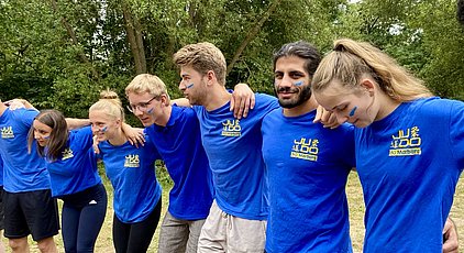 Eine Gruppe von sieben jungen Erwachsenen steht in einer Reihe und umarmt sich gegenseitig. Alle tragen blaue T-Shirts mit dem Aufdruck „Judo BG Marburg“. Einige haben blaue Streifen auf ihren Wangen und wirken fröhlich und motiviert, während sie gemeinsam in die Kamera blicken.