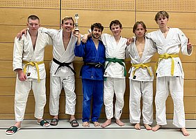Fünf Judoka stehen zusammen in einem Sportraum. Sie tragen verschiedene Judokleidung: einer in Blau, die anderen in Weiß mit farbigen Gürteln (schwarz, grün und gelb). In der Mitte hält ein Kämpfer einen Pokal hoch. Alle lächeln und zeigen eine siegesbewusste Haltung.