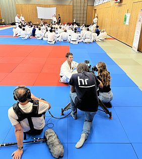 Ein junger Mann im Judogi sitzt auf einer blauen Matte, während er von einem Kamerateam interviewt wird. Im Hintergrund sind viele andere Judokas in weißen Anzügen versammelt. Die Szenerie vermittelt eine sportliche Atmosphäre während eines Judotrainings oder -events.