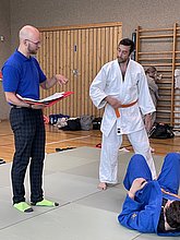 Ein Trainer in blauer Kleidung steht mit einem Notizbuch und spricht zu einem Judoka in einer weißen Uniform mit orangefarbenem Gürtel. Im Hintergrund sind weitere Personen und Trainingsmatten sichtbar. Zwei weitere Judokas liegen auf den Matten.