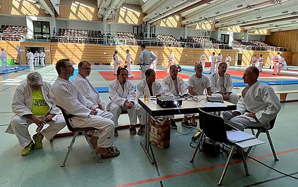 Eine Gruppe von Judokas in weißen Anzügen sitzt an einem Tisch in einer Sporthalle. Einige Teilnehmer führen im Hintergrund Trainingseinheiten aus. Ein Mann spricht mit einem Projektor, während die anderen zuhören. Die Atmosphäre wirkt entspannt und gemeinschaftlich.
