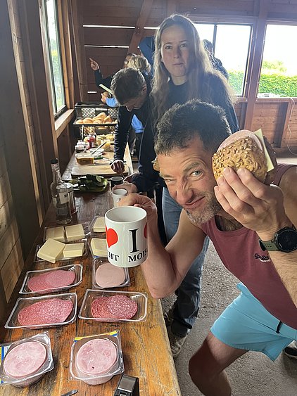 Eine Gruppe von Menschen steht in einem Raum mit einer Holztheke, die mit verschiedenen Lebensmitteln bedeckt ist, darunter Aufschnitt und Käse. Ein Mann mit einem Brötchen in der Hand lächelt fröhlich, während eine Frau im Hintergrund auf etwas zeigt.