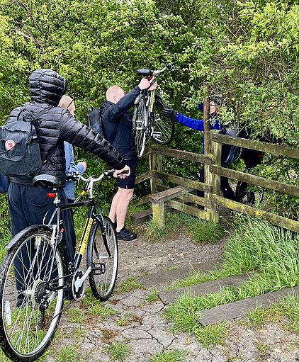 Eine Gruppe von Radfahrern steht an einem Zaun. Einer hebt sein Fahrrad über den Zaun, während ein anderer hilft. Im Hintergrund sind grüne Sträucher und Bäume zu sehen. Der Boden ist uneben und mit Gras und Erde bedeckt.