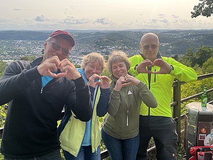 Vier Personen stehen auf einer Aussichtsplattform und formen mit ihren Händen Herzsymbole. Sie lächeln und genießen die Aussicht auf eine grüne Landschaft mit einer Stadt im Hintergrund. Die Stimmung ist freundlich und harmonisch.