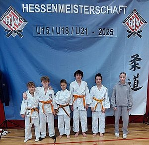 Sechs Kinder in weißen Judoanzügen mit orangefarbener Gürteln stehen vor einem Banner, das "Hessenmeisterschaft U15 / U18 / U21 - 2025" zeigt. Sie lächeln in die Kamera und stehen auf einer Sportbodenfläche.