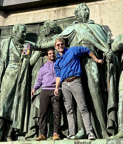 Zwei Männer stehen lächelnd vor einer großen Statue, die historische Figuren darstellt. Der linke Mann trägt einen lila Hoodie, während der rechte Mann einen blauen Hoodie trägt. Beide sind an der Statue angelehnt und scheinen Spaß zu haben.