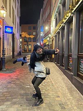 Eine Frau trägt einen Mann auf ihrem Rücken, während sie auf einer gepflasterten Straße stehen. Beide lächeln und scheinen Spaß zu haben. Die Umgebung ist beleuchtet und zeigt einige Geschäfte und eine Straßenlaterne im Hintergrund. Es ist Nacht.