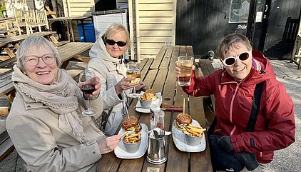 Drei Frauen sitzen an einem Holztisch im Freien und halten Getränkegläser in der Hand. Vor ihnen stehen Teller mit Burgern und Pommes. Es herrscht eine fröhliche Atmosphäre, und alle scheinen den Moment zu genießen.