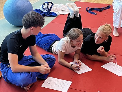 Drei Jugendliche sitzen auf einer roten Matte und diskutieren. Zwei von ihnen liegen auf der Seite und halten Stifte sowie Blätter mit Notizen. Der dritte Jugendliche sitzt und hört aufmerksam zu. Im Hintergrund ist ein blauer Gymnastikball zu sehen.
