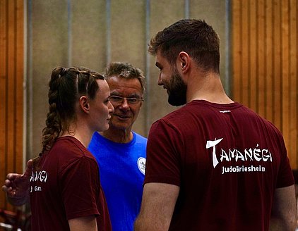 Drei Personen unterhalten sich in einer Sporthalle. Eine Frau mit geflochtenem Haar trägt ein rotes T-Shirt, während ein Mann mit Bart eine ähnliche Farbgebung hat. Im Hintergrund steht ein älterer Mann in einem blauen Shirt, der lächelt und die beiden anspricht.