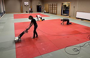 Mehrere Personen reinigen einen Sportboden in einem großen Raum. Eine Person benutzt eine Maschine, um den roten Boden zu säubern, während zwei andere auf dem Boden knien und reinigen. Im Hintergrund sind Sportgeräte und eine Basketballkorb zu sehen.