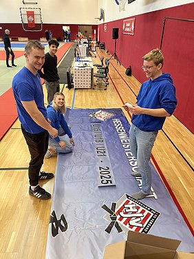 Eine Gruppe junger Leute bereitet ein Banner für eine Meisterschaft vor. Sie lächeln und arbeiten gemeinsam auf dem Boden in einer Sporthalle. Das Banner zeigt den Schriftzug „Hessische Meisterschaft U8/U21 - 2025“.