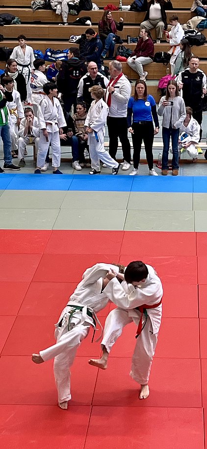 Zwei Kinder in Judokleidung kämpfen auf einer roten Matte. Einer der Gegner führt einen Wurf aus, während der andere versucht, sich zu verteidigen. Im Hintergrund sind Zuschauer zu sehen, die das Geschehen beobachten.